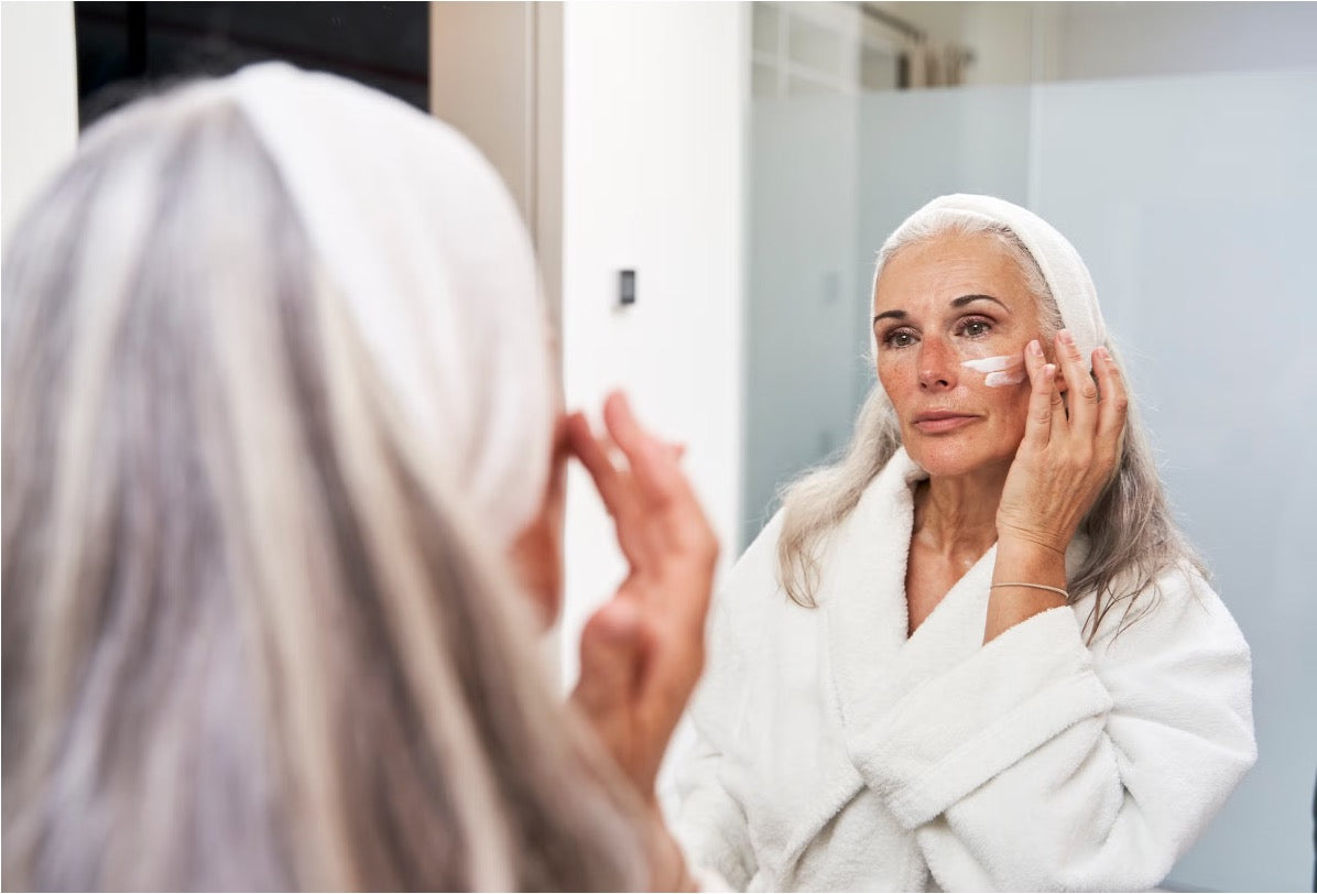 Older woman in a white bathrobe applying cream in a bathroom mirror as part of her anti-aging skincare routine