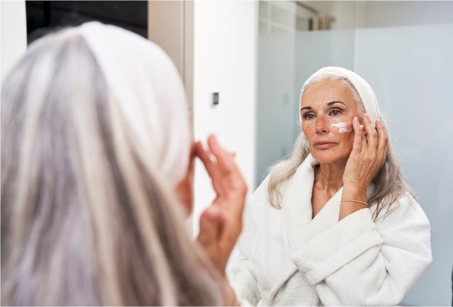 Older woman in a white bathrobe applying cream in a bathroom mirror as part of her anti-aging skincare routine