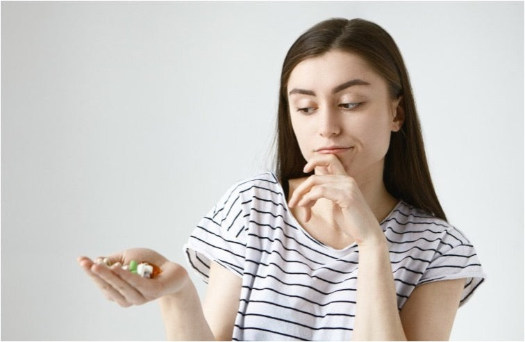 Young woman in a striped shirt looking thoughtfully at a handful of assorted pills and capsules